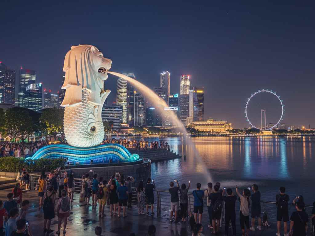 Patung Merlion di Singapura yang menyemburkan air dengan latar belakang gemerlap lampu kota dan Singapore Flyer di malam hari.