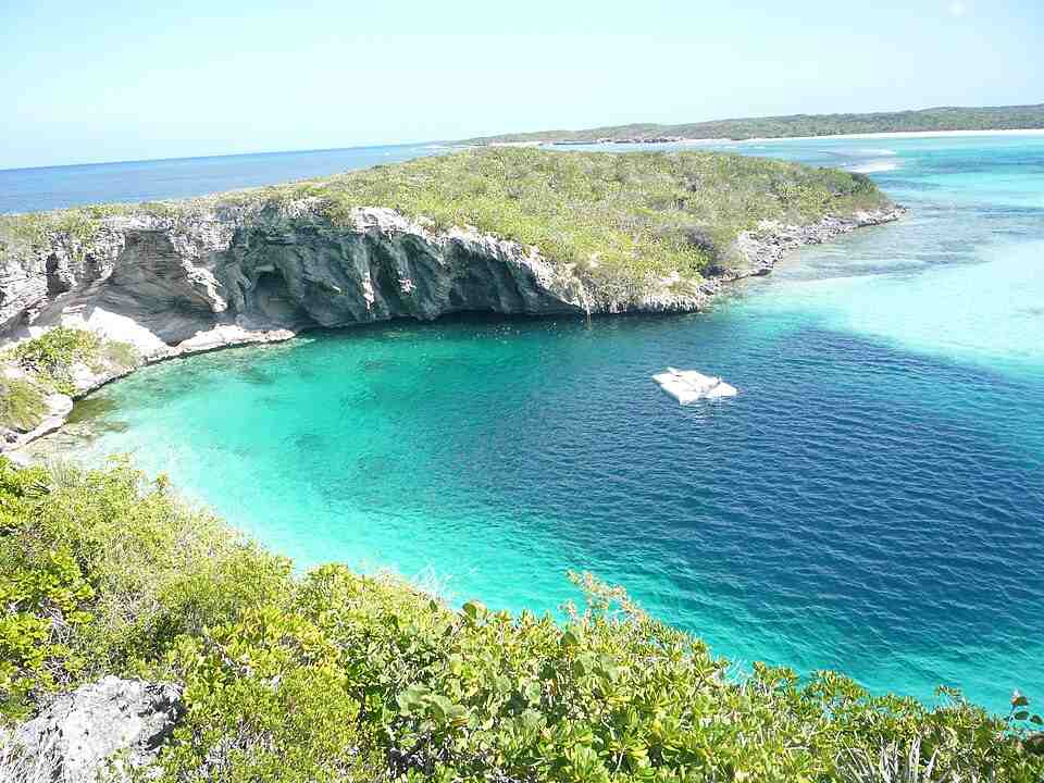 Foto Dean’s Blue Hole di Bahama menampilkan kolam alami berbentuk bundar dengan air biru tua di tengah pantai karang, dikelilingi tebing dan vegetasi tropis.