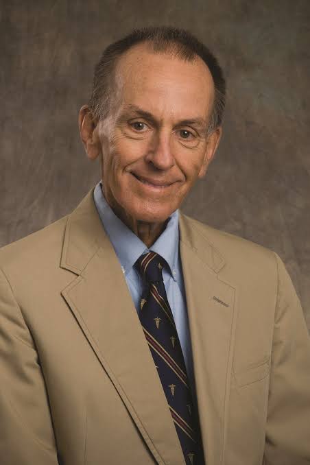 Potret Dr. John Seidensticker mengenakan jas cokelat dan dasi biru dalam latar belakang studio.