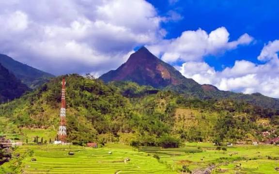 Pemandangan perbukitan hijau dengan sistem sawah terasering yang menghadap ke puncak Gunung Muria yang lancip.