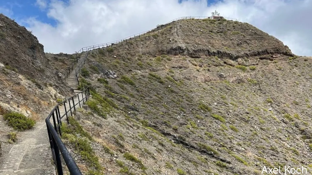 Jalur pendakian batu curam menuju puncak Kelimutu, Nusa Tenggara Timur, dilengkapi dengan pagar pembatas besi hitam di lereng bukit vulkanik yang tandus.