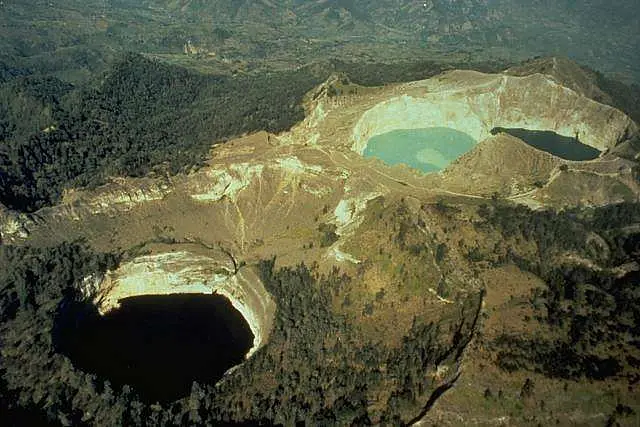 Pemandangan udara Gunung Kelimutu menunjukkan tiga danau kawah yang berbeda. Salah satu danau berwarna biru muda kehijauan, yang lain berwarna hijau gelap, dan yang ketiga berwarna hitam pekat, dikelilingi oleh hutan hijau lebat dan lereng vulkanik.