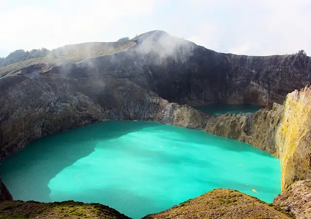 Pemandangan close-up Danau Kelimutu, menunjukkan air kawah berwarna biru kehijauan cerah (aqua) yang kontras dengan dinding kawah curam berwarna cokelat gelap dan hijau.